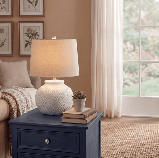 A table lamp with a textured white ceramic base and a beige fabric drum shade sits on a navy blue nightstand in a cozy, warm-toned living room setting.