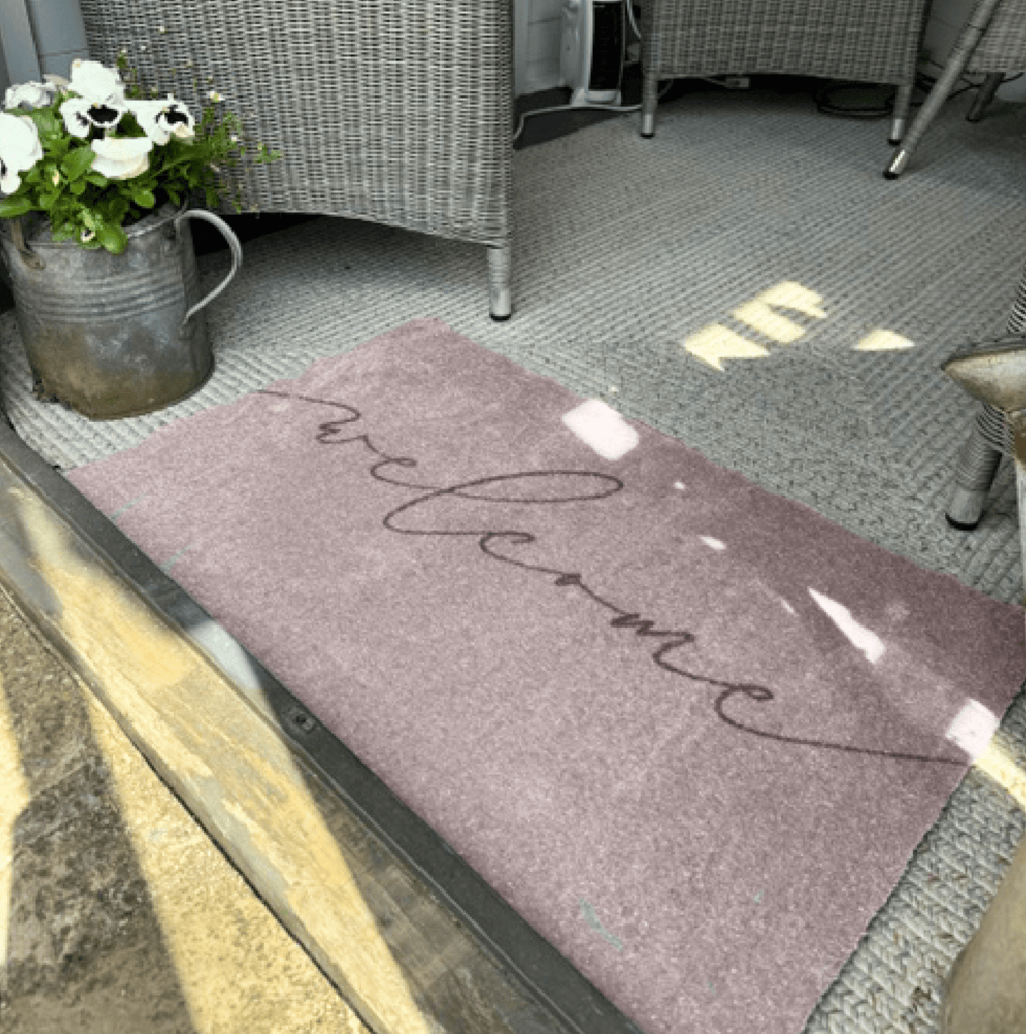 Grey welcome mat on a patio with a metal watering can and flowers.
