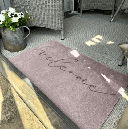 Grey welcome mat on a patio with a metal watering can and flowers.