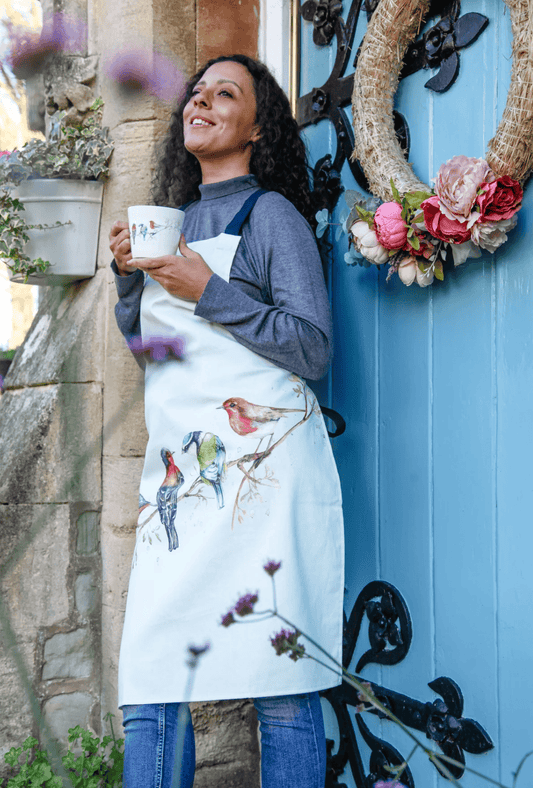 Woman wearing a white apron with bird designs, holding a mug, standing in front of a blue door with a wreath.