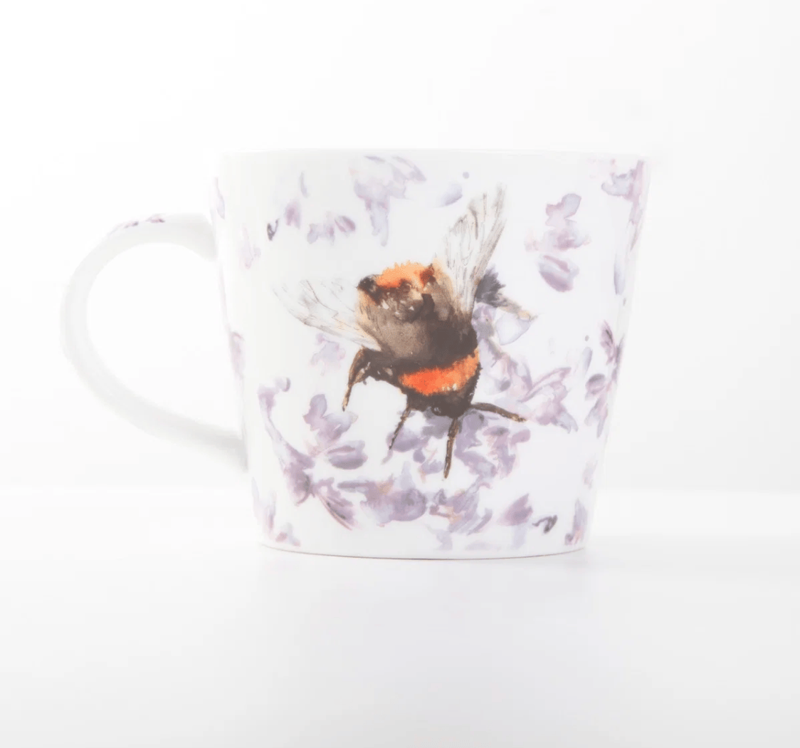 A white ceramic mug featuring a watercolor illustration of a bumblebee over light purple flowers, set against a clean white background.