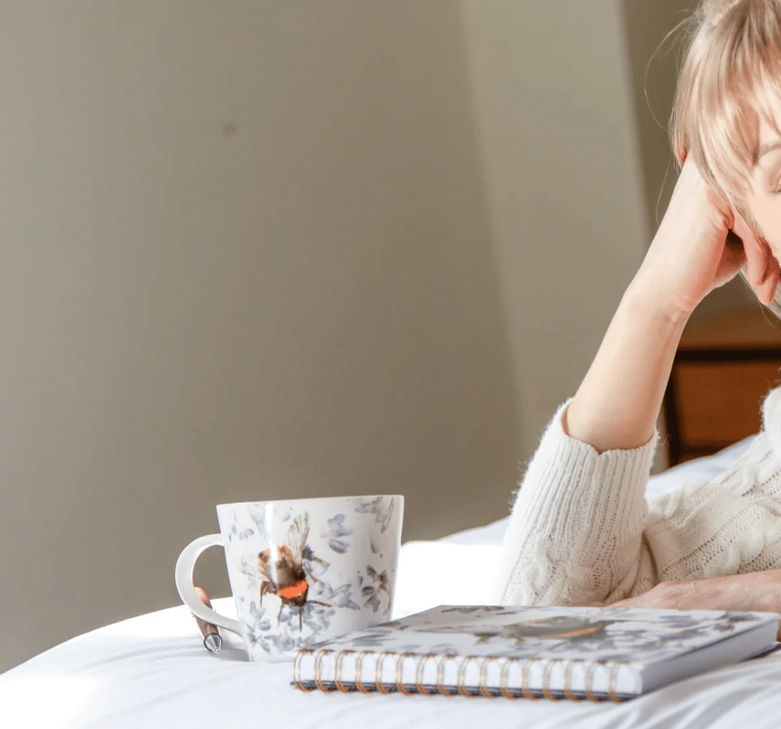 A woman in a cream knit sweater rests her head on her hand on a bed, with a white floral mug and a spiral notebook in the foreground.