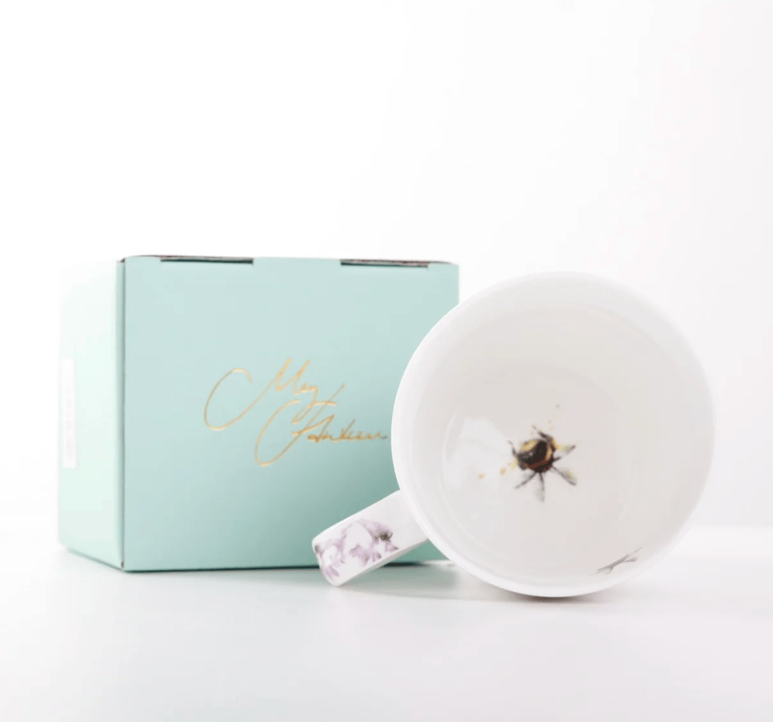 A white ceramic mug with a floral-patterned handle and a printed bee design inside the cup, shown next to its light blue gift box with gold script lettering on a clean white background.