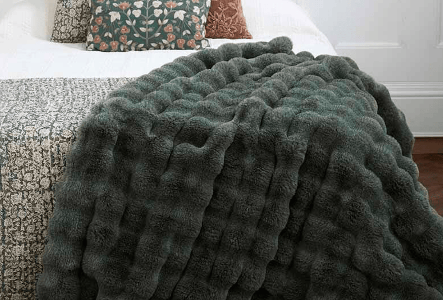 A chunky, ribbed plush throw blanket in a dark sage green color draped over a bed with a patterned quilt and decorative pillows in a bedroom setting.