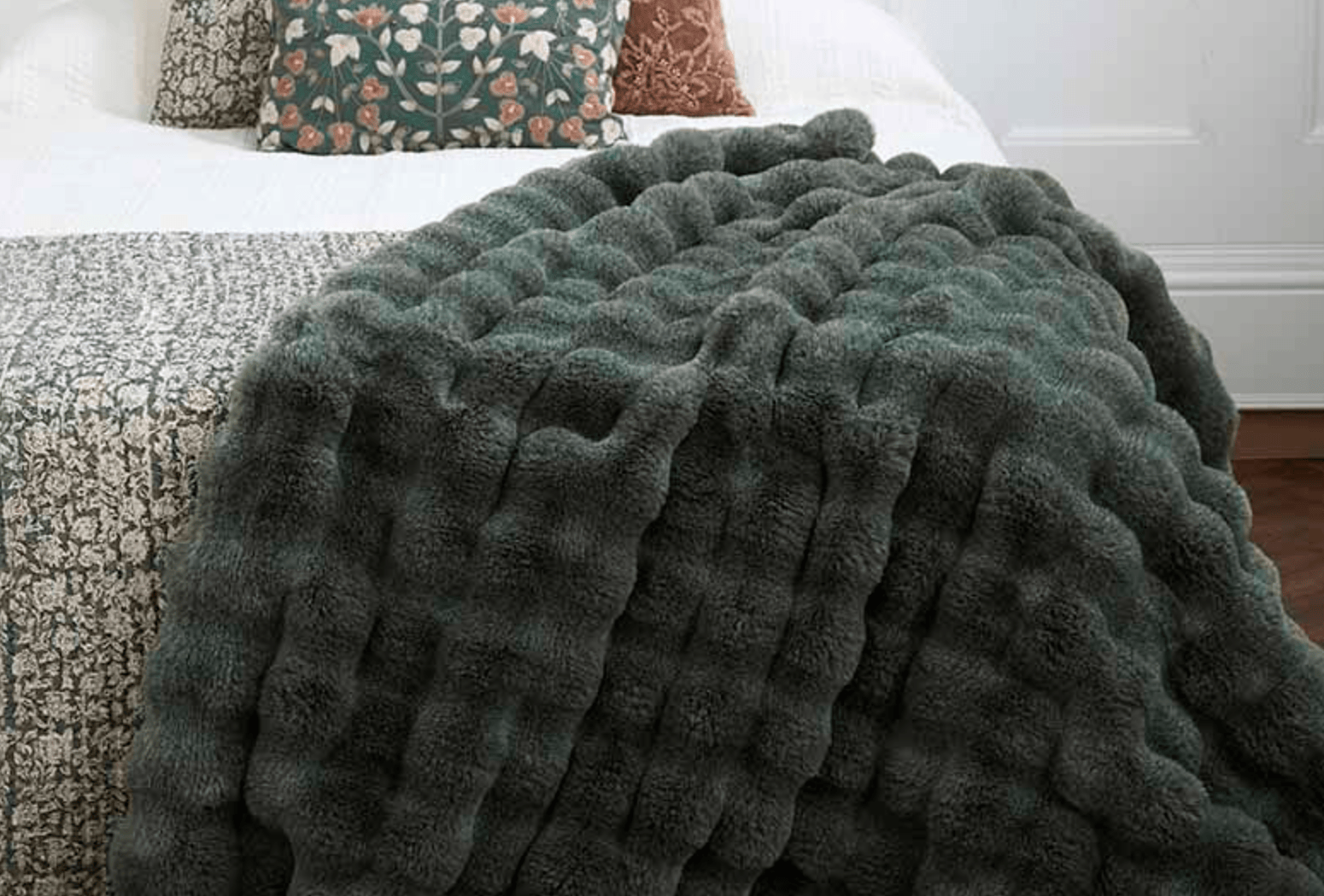 A chunky, ribbed plush throw blanket in a dark sage green color draped over a bed with a patterned quilt and decorative pillows in a bedroom setting.