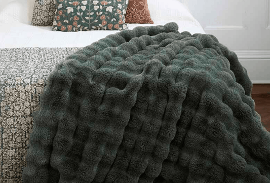 A chunky, ribbed plush throw blanket in a dark sage green color draped over a bed with a patterned quilt and decorative pillows in a bedroom setting.