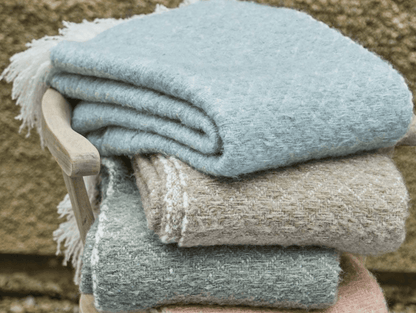 Stack of folded blankets in various colors on a wooden stool against a stone wall.