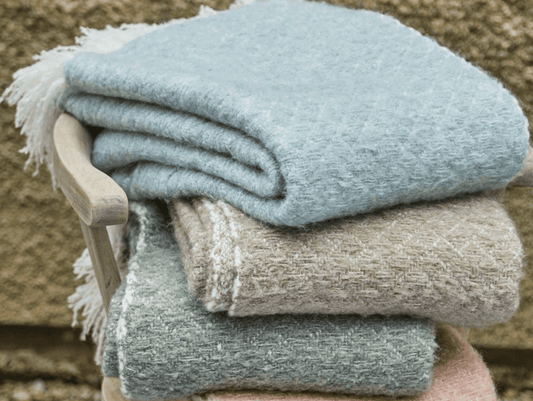 Stack of folded blankets in various colors on a wooden stool against a stone wall.