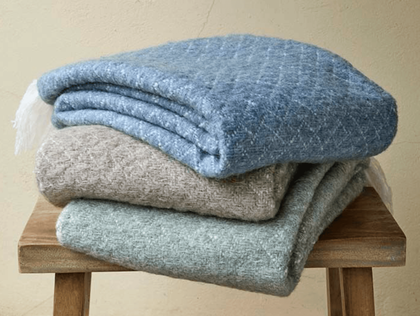 Stack of three folded blankets in blue, gray, and green on a wooden stool against a beige wall.