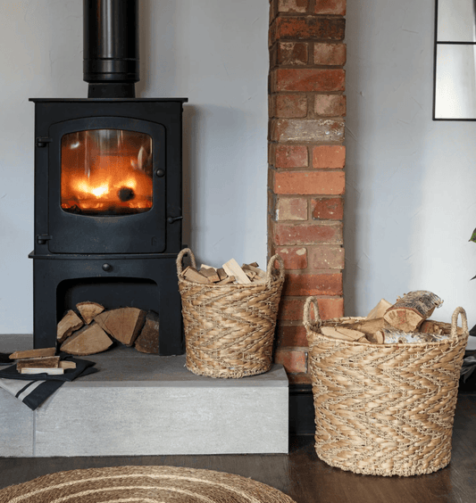 Wood-burning stove with fire lit, surrounded by baskets of wood logs against a brick wall.
