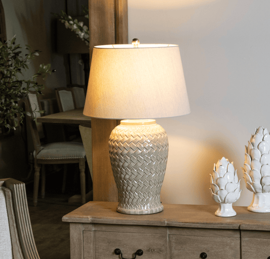 A table lamp with a textured cream ceramic base and a beige fabric drum shade, glowing with a warm light, styled on a wooden console table in a dining room setting.